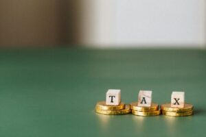 A green table fades from the foreground into the background with three gold coins sitting on the right hand side. Each coin features a little wooden block, and the blocks have a letter that spells out tax,